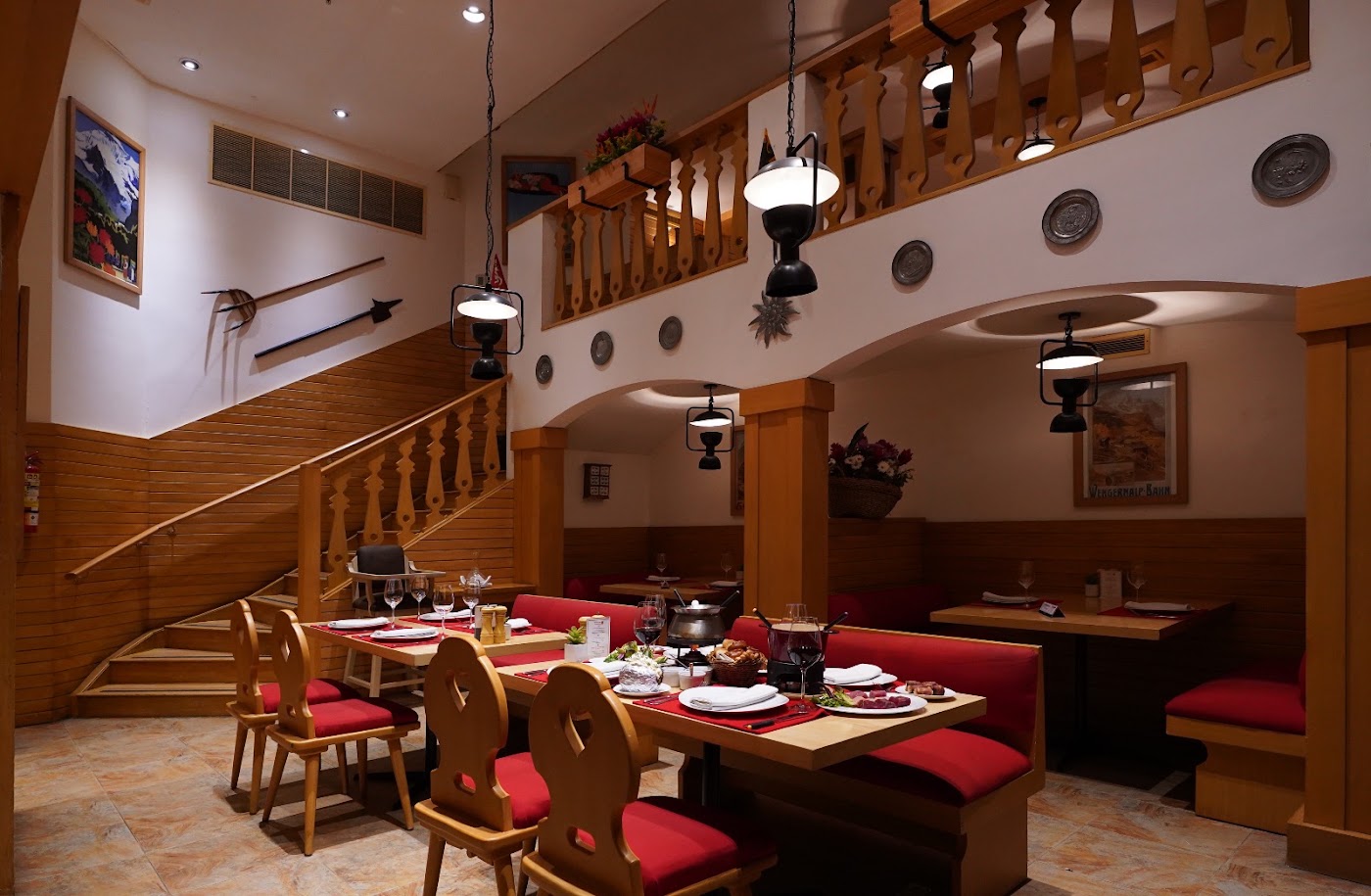 Salón de La Fondue con mesas de madera, asientos rojos, lámparas colgantes y una escalera al nivel superior